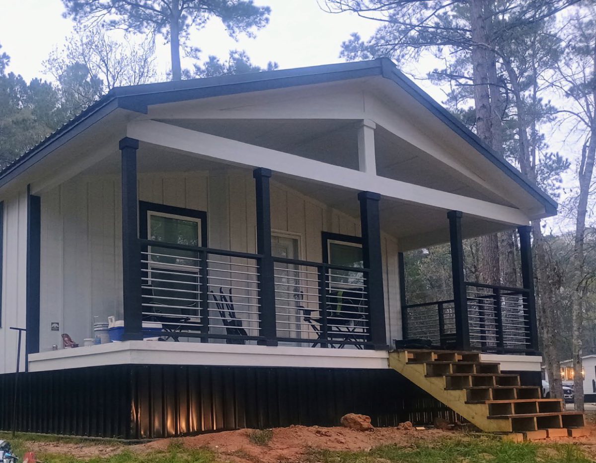 The front of a manufactured home with a deep covered porch, navy posts and dark skirting, on a green lot under pines.