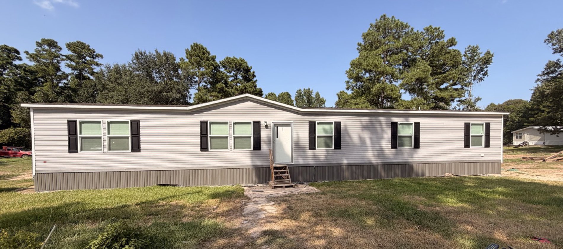 A long cream manufactured home with black shutters, finished with skirting that runs clean along the ground, on a mown green lot backed by pines.