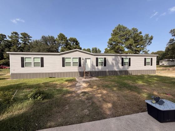 A long cream manufactured home with black shutters, skirted the full length, on a mown green lot.